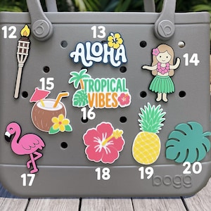 May include: A gray rubber tote bag featuring nine colorful decorative charms numbered 12 through 20. The charms include a tiki torch, the text ALOHA, a hula dancer, a coconut drink, TROPICAL VIBES text, a flamingo, a hibiscus, a pineapple, and a leaf.