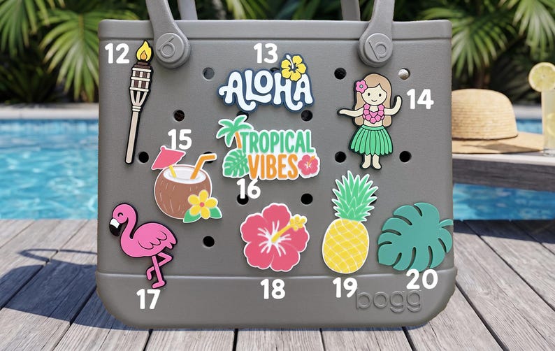 May include: A gray rubber tote bag featuring nine colorful decorative charms numbered 12 through 20. The charms include a tiki torch, the text ALOHA, a hula dancer, a coconut drink, TROPICAL VIBES text, a flamingo, a hibiscus, a pineapple, and a leaf.