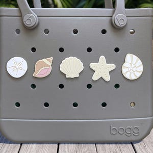 May include: A gray Bogg tote bag featuring a perforated design with five decorative charms attached to the front. The charms include a white sand dollar, a pink and tan conch shell, a cream scallop shell, a beige starfish, and a white nautilus shell.