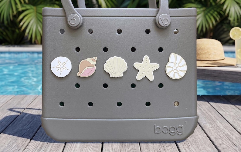 May include: A gray Bogg tote bag featuring a perforated design with five decorative charms attached to the front. The charms include a white sand dollar, a pink and tan conch shell, a cream scallop shell, a beige starfish, and a white nautilus shell.