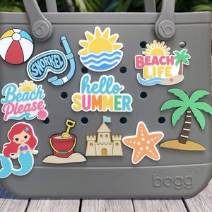 May include: A gray Bogg bag featuring various summer-themed charms including a beach ball, snorkel, sun, palm tree, mermaid, sandcastle, starfish, and bucket. The charms display text such as Snorkel, Beach Life, Beach Please, and Hello Summer.