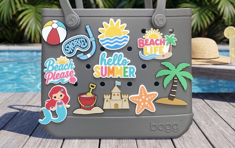 May include: A gray Bogg bag featuring various summer-themed charms including a beach ball, snorkel, sun, palm tree, mermaid, sandcastle, starfish, and bucket. The charms display text such as Snorkel, Beach Life, Beach Please, and Hello Summer.