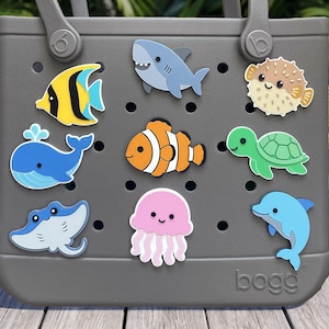 May include: A gray tote bag featuring a series of holes decorated with various colorful sea creature charms, including a yellow angelfish, a blue whale, a gray shark, an orange clownfish, a pink jellyfish, a green sea turtle, and a blue dolphin.
