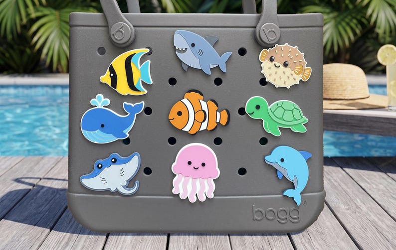 May include: A gray tote bag featuring a series of holes decorated with various colorful sea creature charms, including a yellow angelfish, a blue whale, a gray shark, an orange clownfish, a pink jellyfish, a green sea turtle, and a blue dolphin.