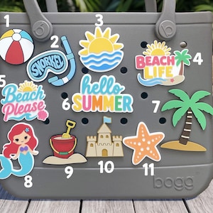 May include: A gray tote bag featuring eleven colorful summer-themed decorative inserts. The inserts include a beach ball, snorkel, sun, beach life text, beach please text, hello summer text, palm tree, mermaid, bucket, sandcastle, and starfish.
