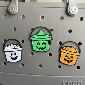 May include: Three Halloween-themed charms in the shape of buckets with black handles. One is white, one is green with a witch hat, and one is orange with a jack-o'-lantern face. The charms are attached to a gray bag.