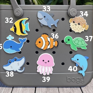 May include: A gray rubber tote bag featuring nine colorful sea creature charms attached to its perforated surface. The charms include a yellow fish, shark, pufferfish, blue whale, clownfish, sea turtle, stingray, jellyfish, and dolphin, labeled 32-40.