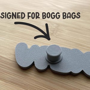 May include: A gray, textured, custom-shaped accessory featuring a central cylindrical post on the back. The text "DESIGNED FOR BOGG BAGS" is printed above the object with a black arrow pointing downward toward the center of the accessory.