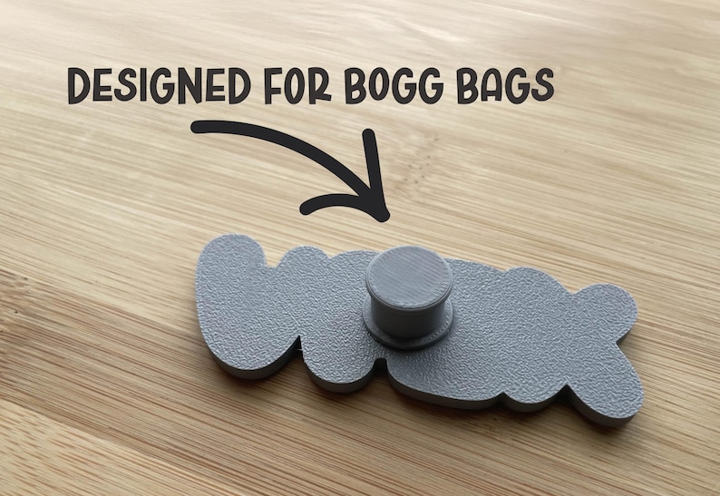 May include: A gray, textured, custom-shaped accessory featuring a central cylindrical post on the back. The text "DESIGNED FOR BOGG BAGS" is printed above the object with a black arrow pointing downward toward the center of the accessory.