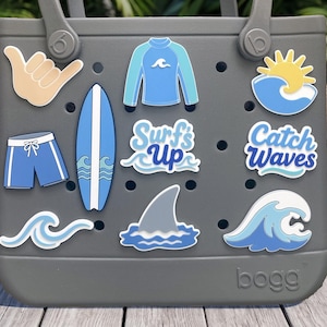 May include: A gray tote bag featuring various surf-themed charms including a shaka hand sign, a blue rash guard, a yellow sun over a wave, blue swim trunks, a surfboard, a shark fin, and text charms that read Surf's Up and Catch Waves.
