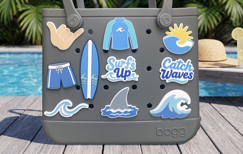 May include: A gray tote bag featuring various surf-themed charms including a shaka hand sign, a blue rash guard, a yellow sun over a wave, blue swim trunks, a surfboard, a shark fin, and text charms that read Surf's Up and Catch Waves.