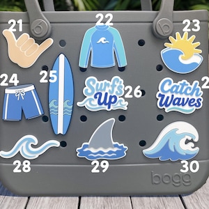 May include: A gray tote bag featuring ten decorative charms with surfing themes, including a shaka hand sign, a rash guard, a sun over a wave, swim trunks, a surfboard, text reading Surf's Up, text reading Catch Waves, and various wave and shark fin icons.