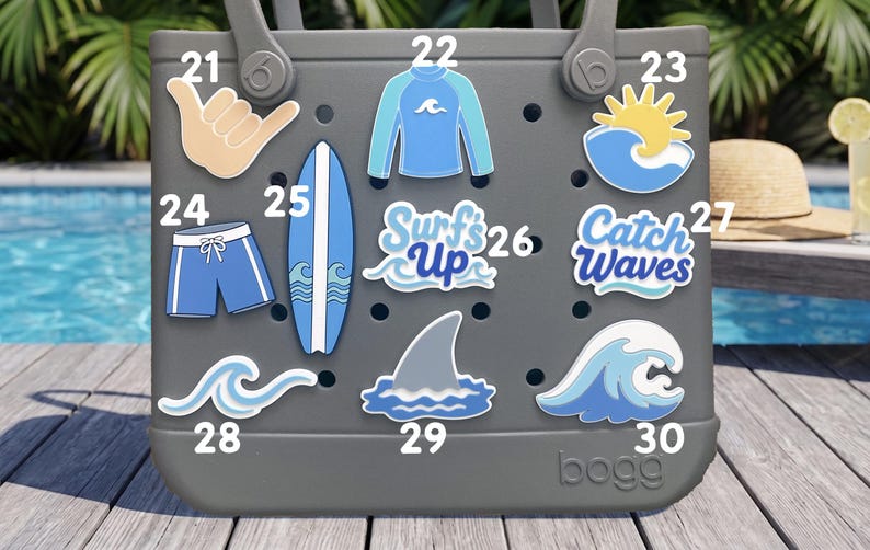 May include: A gray tote bag featuring ten decorative charms with surfing themes, including a shaka hand sign, a rash guard, a sun over a wave, swim trunks, a surfboard, text reading Surf's Up, text reading Catch Waves, and various wave and shark fin icons.