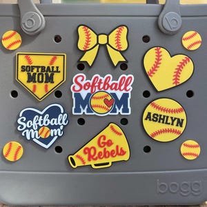 May include: Gray tote bag decorated with softball-themed charms. Charms include a yellow bow, hearts, softballs, and text that says "Softball Mom", "Ashlyn", and "Go Rebels". A softball is visible on the left.