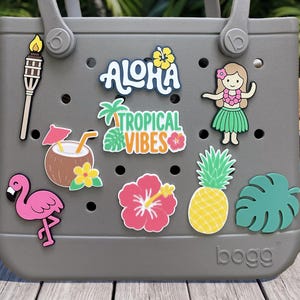 May include: A gray tote bag with circular ventilation holes, decorated with various tropical-themed charms including a tiki torch, a coconut drink, a pink flamingo, a pineapple, a hibiscus flower, a hula dancer, and text reading ALOHA TROPICAL VIBES.