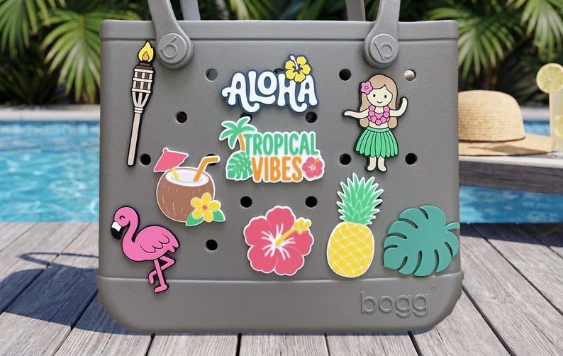 May include: A gray tote bag with circular ventilation holes, decorated with various tropical-themed charms including a tiki torch, a coconut drink, a pink flamingo, a pineapple, a hibiscus flower, a hula dancer, and text reading ALOHA TROPICAL VIBES.