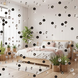 May include: A bedroom scene with a bed, plants, and a decorative curtain of hanging silver and black spheres. The bed has white bedding and pink pillows. Potted plants are placed around the room, and the curtain adds a touch of sparkle.
