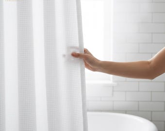 Puede incluir: Cortina de ducha blanca con tejido de gofre. Una mano sostiene la cortina abierta, revelando una bañera blanca y una ventana. La cortina tiene un patrón texturizado en forma de rejilla. El baño tiene azulejos blancos.