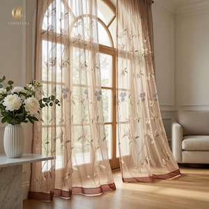 Op de afbeelding: Transparante beige gordijnen met delicate geborduurde bloemmotieven in zachte roze, blauwe en groene tinten. De gordijnen hebben een effen donkerbruine rand aan de onderkant en hangen aan een roede voor een groot boogvenster met houten kozijnen.