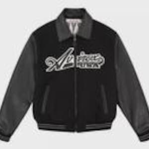 K&ouml;nnte beinhalten: Eine schwarze Collegejacke mit langen schwarzen &Auml;rmeln, einem dunkelgrauen Kragen und einem Frontrei&szlig;verschluss. Auf der Brust befindet sich eine wei&szlig;e Stickerei mit dem Schriftzug "Avirex USA" in kursiver Schrift. Saum und &Auml;rmelb&uuml;ndchen sind gestreift.
