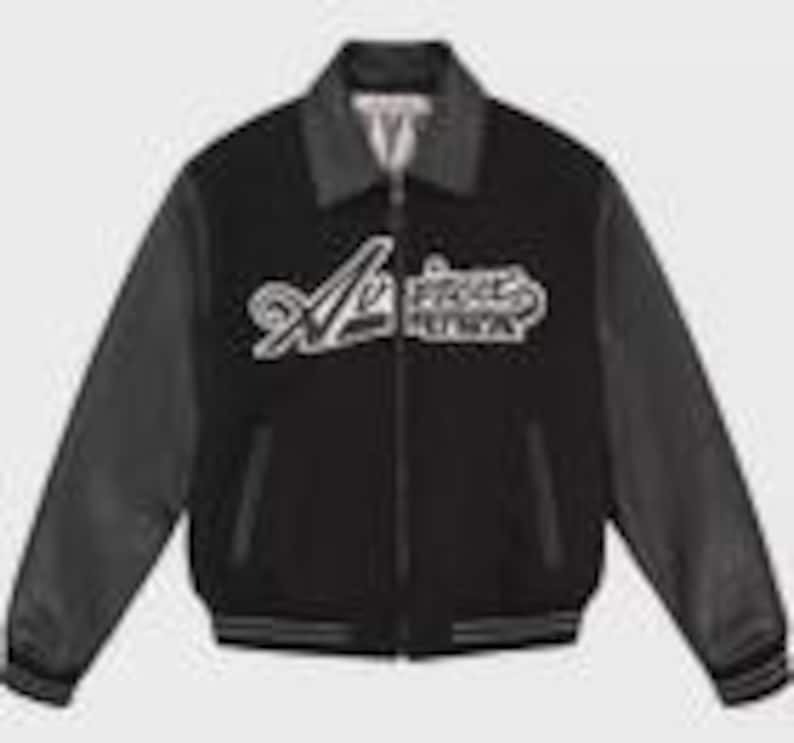 K&ouml;nnte beinhalten: Eine schwarze Collegejacke mit langen schwarzen &Auml;rmeln, einem dunkelgrauen Kragen und einem Frontrei&szlig;verschluss. Auf der Brust befindet sich eine wei&szlig;e Stickerei mit dem Schriftzug "Avirex USA" in kursiver Schrift. Saum und &Auml;rmelb&uuml;ndchen sind gestreift.