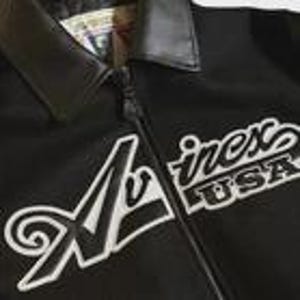 K&ouml;nnte beinhalten: Eine schwarze Jacke mit dunklem Kragen und Rei&szlig;verschluss auf der Vorderseite. Auf der Brust befindet sich eine wei&szlig;e Stickerei mit der Aufschrift Avirex USA und einem gro&szlig;en, stilisierten Buchstaben A. Der Stoff wirkt glatt und einfarbig schwarz.