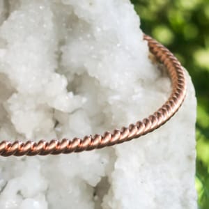 Könnte beinhalten: Ein kupferfarbenes, gedrehtes Drahtarmband. Das Armband befindet sich vor einem weißen, strukturierten Hintergrund mit grünem Laub im Hintergrund. Das Design des Armbands weist eine Reihe eng gewickelter, paralleler Stränge auf.