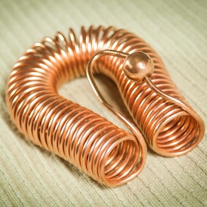 Puede incluir: Una bobina de cobre con un acento esférico. La bobina es de un color cobre brillante y brillante y está enrollada en forma de espiral. El acento esférico también es de cobre. El objeto descansa sobre una superficie texturizada verde claro.