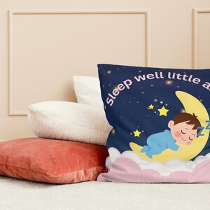 Op de afbeelding: Een decoratief kussen met een marineblauwe achtergrond met een slapende baby op een maan. Het kussen heeft de tekst "sleep well little angel" en is omgeven door sterren en wolken. Andere kussens zijn op de achtergrond te zien.