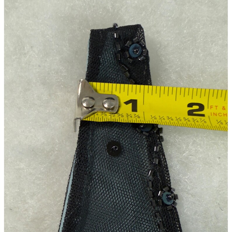 May include: Close-up of a black mesh fabric with blue accents and embellishments. The fabric is measured with a yellow tape measure, showing a length of approximately 1.5 inches. The fabric is decorated with small beads and sequins.
