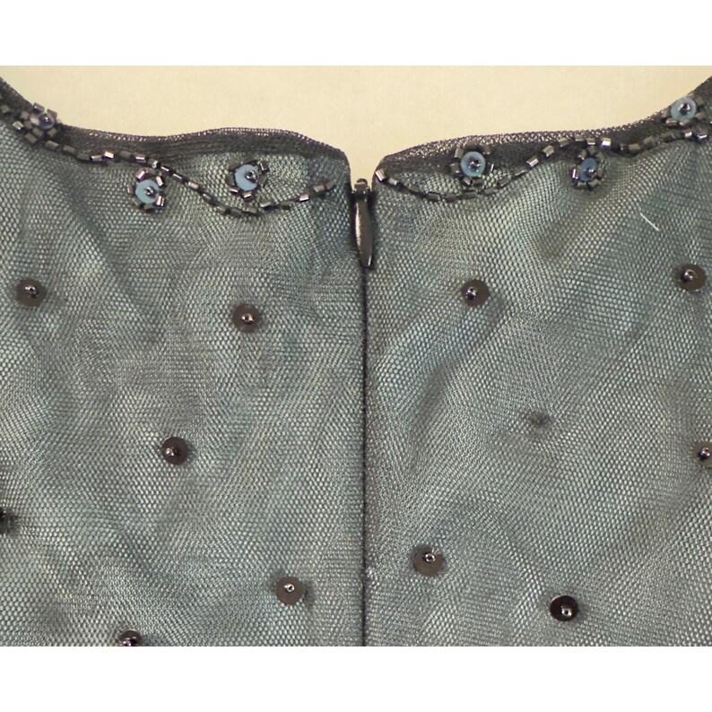 May include: Close-up of a teal-colored garment with a mesh overlay. The neckline is embellished with small, light blue beads and silver beading. Scattered, dark gray sequins are sewn onto the mesh fabric. A silver zipper runs down the center.