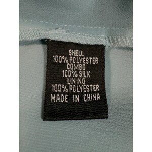 May include: A close-up of a black tag with white text. The tag reads "SHELL 100% POLYESTER COMBO 100% SILK LINING 100% POLYESTER MADE IN CHINA" against a light blue fabric background.