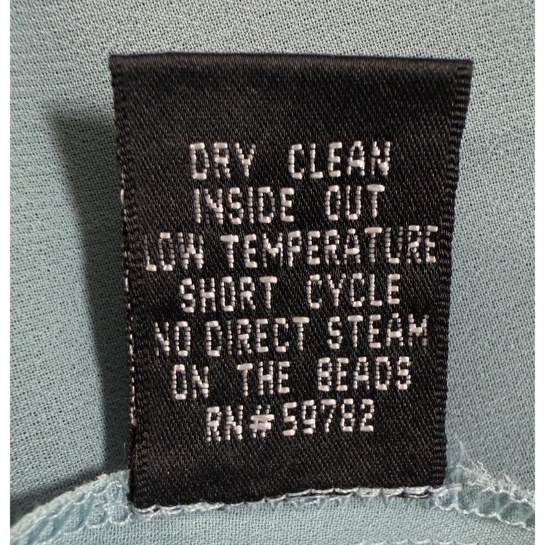 May include: A black clothing tag with white text, including care instructions: "DRY CLEAN INSIDE OUT LOW TEMPERATURE SHORT CYCLE NO DIRECT STEAM ON THE BEADS RN#59782". The tag is against a light blue fabric.