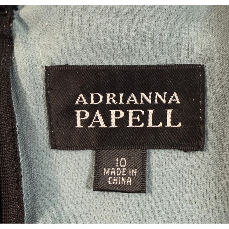 May include: Close-up of a black label with the text "ADRIANNA PAPELL" in white. Below the main label is a smaller label that reads "10 MADE IN CHINA". The background is a light blue fabric.
