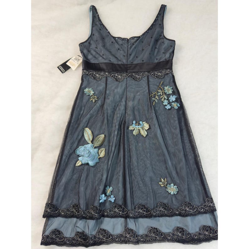 May include: A sleeveless, knee-length dress in shades of blue and black. The dress features a V-neck, a black satin ribbon at the waist, and floral embroidery in shades of blue and green. The skirt has a layered design with a black lace trim.