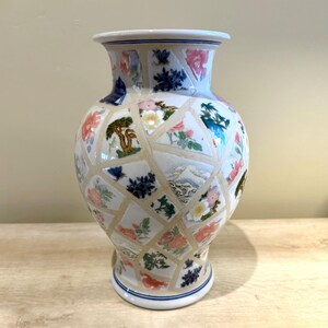 May include: A decorative ceramic vase with a mosaic-like design. The vase features a white base with a pattern of floral and landscape motifs in various colors, including pink, blue, and green. The vase has a wide body and a narrow neck.