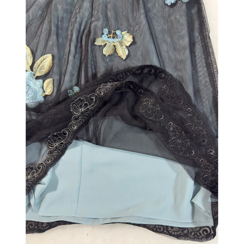 May include: A black tulle skirt with floral embroidery in shades of blue and gold. The skirt features a light blue lining and is trimmed with black lace. The delicate design suggests a formal or special occasion garment.