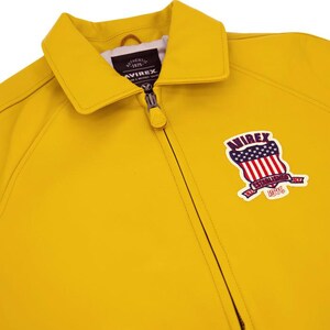 Puede incluir: Chaqueta amarilla brillante con cierre de cremallera y cuello cl&aacute;sico. La chaqueta presenta un parche en la parte delantera con la palabra "AVIREX", un dise&ntilde;o de escudo y el texto "ESTABLISHED 1975".