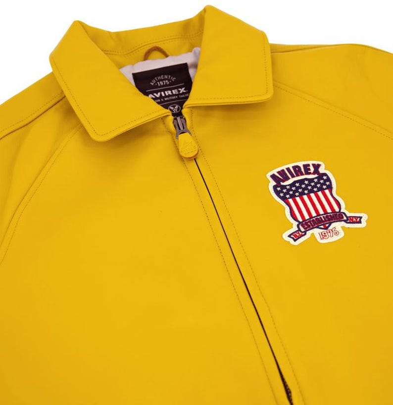 Puede incluir: Chaqueta amarilla brillante con cierre de cremallera y cuello cl&aacute;sico. La chaqueta presenta un parche en la parte delantera con la palabra "AVIREX", un dise&ntilde;o de escudo y el texto "ESTABLISHED 1975".
