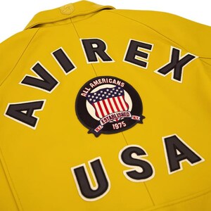 Puede incluir: Primer plano de una chaqueta amarilla con la palabra "AVIREX" y "USA" en letras negras. Tambi&eacute;n es visible un parche circular con la bandera estadounidense y las palabras "ALL AMERICANS ESTABLISHED 1975 N.Y.".