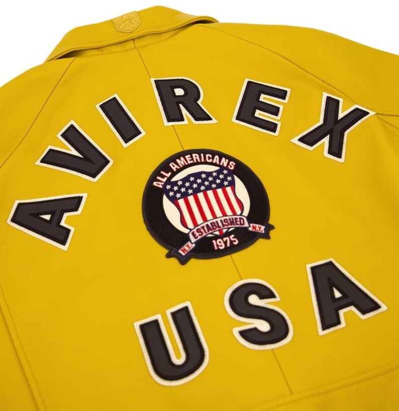 Puede incluir: Primer plano de una chaqueta amarilla con la palabra "AVIREX" y "USA" en letras negras. Tambi&eacute;n es visible un parche circular con la bandera estadounidense y las palabras "ALL AMERICANS ESTABLISHED 1975 N.Y.".