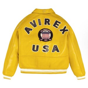 Puede incluir: Chaqueta de cuero amarillo brillante con la palabra "AVIREX" arqueada en la parte superior de la espalda. Debajo, se muestra la palabra "USA". Un parche circular con las palabras "ALL AMERICANS ESTABLISHED 1975" y un dise&ntilde;o de la bandera estadounidense est&aacute; centrado encima del texto "USA". La chaqueta tiene pu&ntilde;os a rayas blancas y amarillas.