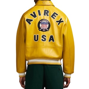 Puede incluir: Chaqueta de cuero amarillo mostaza con la palabra "AVIREX" arqueada sobre los hombros. La parte trasera presenta un parche circular con la bandera estadounidense y las palabras "ALL AMERICANS ESTABLISHED 1975 USA". El dobladillo es a rayas blancas y amarillas.