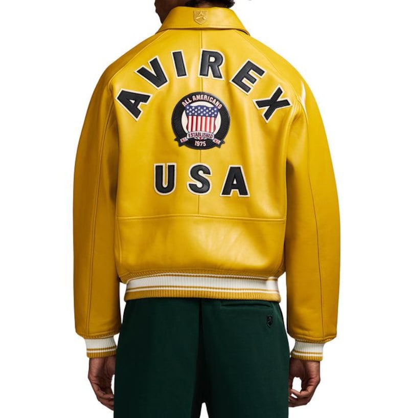 Puede incluir: Chaqueta de cuero amarillo mostaza con la palabra "AVIREX" arqueada sobre los hombros. La parte trasera presenta un parche circular con la bandera estadounidense y las palabras "ALL AMERICANS ESTABLISHED 1975 USA". El dobladillo es a rayas blancas y amarillas.