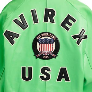 Puede incluir: Chaqueta de cuero verde brillante con letras negras y blancas que deletrean "AVIREX" y "USA". Un parche circular presenta la bandera estadounidense y las palabras "ALL AMERICANS ESTABLISHED 1975 N.Y."