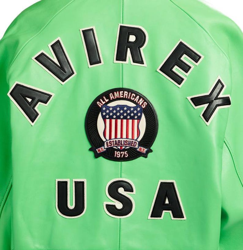 Puede incluir: Chaqueta de cuero verde brillante con letras negras y blancas que deletrean "AVIREX" y "USA". Un parche circular presenta la bandera estadounidense y las palabras "ALL AMERICANS ESTABLISHED 1975 N.Y."