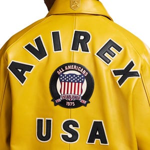 Puede incluir: Chaqueta de cuero amarilla con letras y parches en blanco y negro. La parte trasera de la chaqueta muestra las palabras "AVIREX" y "USA", junto con un parche circular que dice "ALL AMERICANS ESTABLISHED 1975".