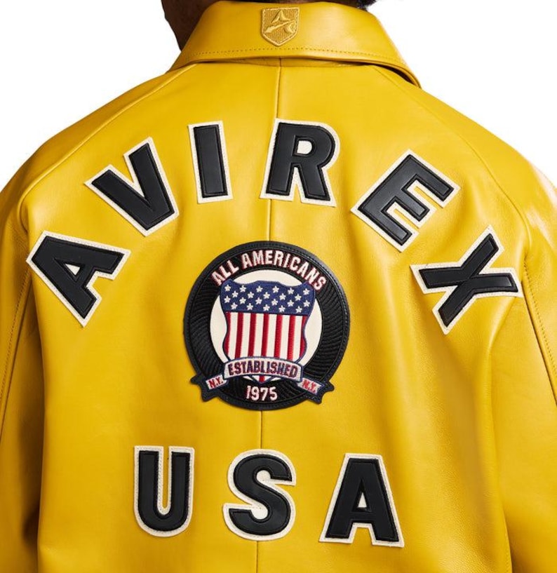 Puede incluir: Chaqueta de cuero amarilla con letras y parches en blanco y negro. La parte trasera de la chaqueta muestra las palabras "AVIREX" y "USA", junto con un parche circular que dice "ALL AMERICANS ESTABLISHED 1975".