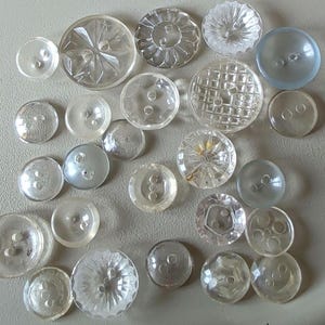 May include: A selection of clear vintage buttons in various sizes and designs. The buttons are made of transparent materials, with faceted, textured, and smooth surfaces. Some have two or four holes for sewing, and others have a shank.