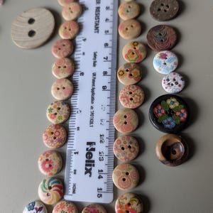 May include: A selection of decorative wooden buttons with floral and patterned designs. The buttons are in various sizes and colours, including pink, red, and green. A ruler shows the buttons are up to 2.5 cm in diameter.